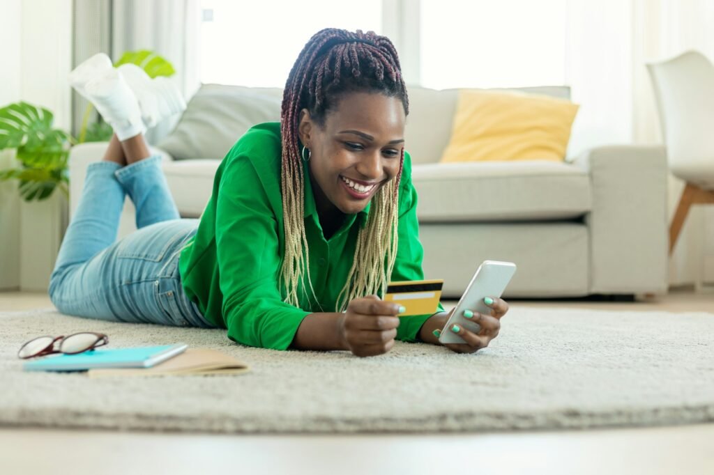 Excited black woman laying on floor carpet with cell phone and plastic credit card, ordering food or